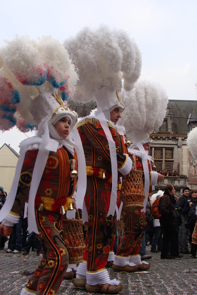 A line of Gilles in white feather hats and costumes with bells and baskets of oranges in a Binche street