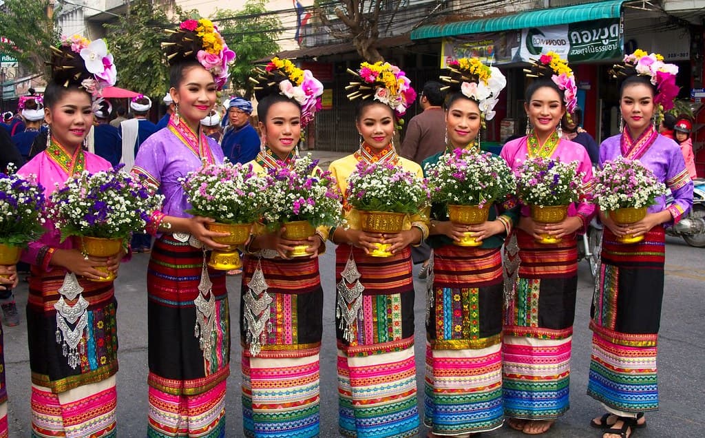 A line of young women in colourful traditional Lanna dress holding flower arrangements wait to join the parade in Chiang Mai