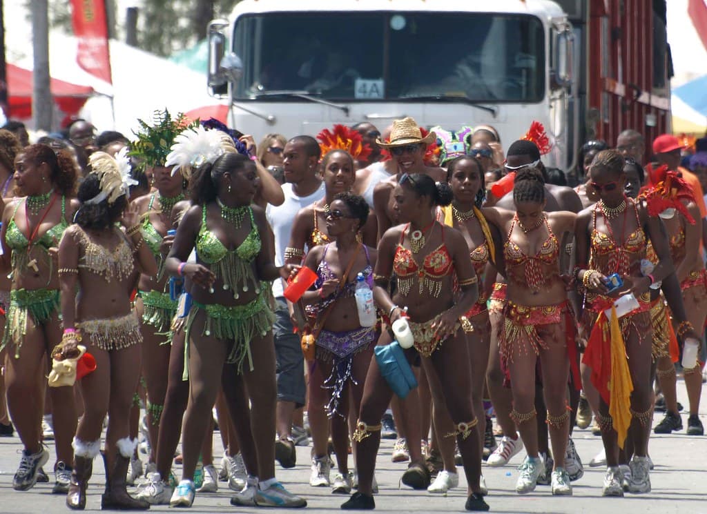 Masqueraders in sequinned costumes and feathered headdresses walk down a road in the Grand Kadooment parade, followed by a music truck