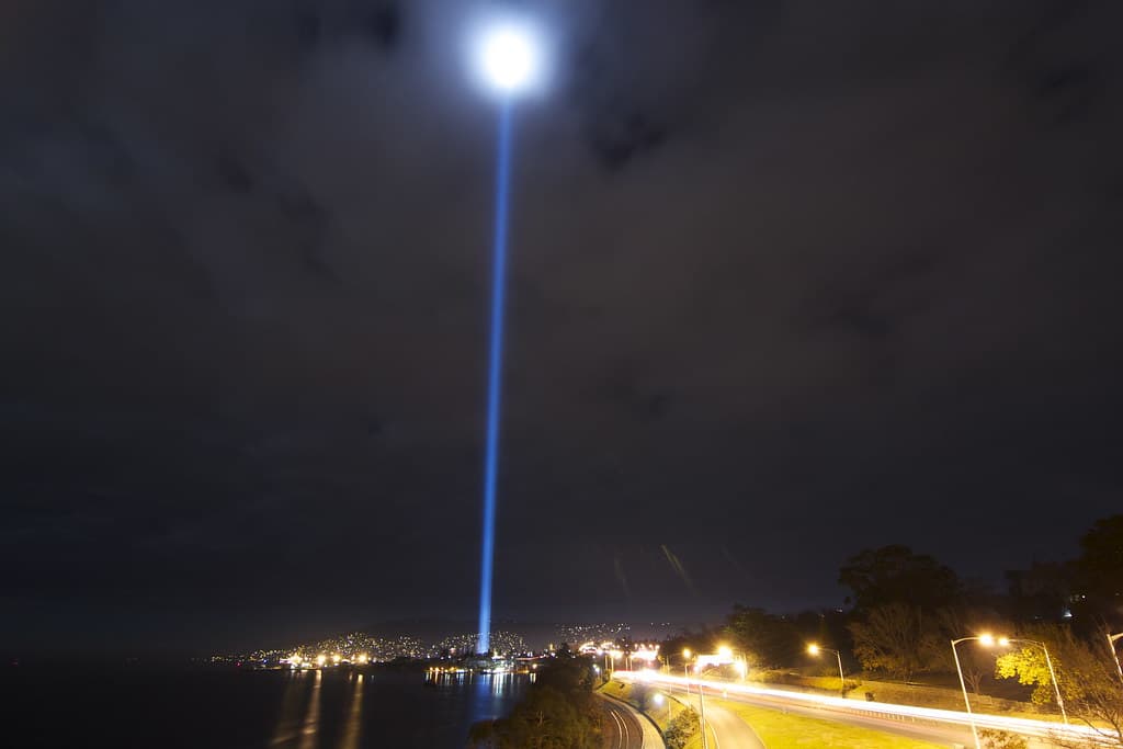 A vertical beam of light rises into the night sky from the waterfront, seen from the Tasman Bridge in Hobart