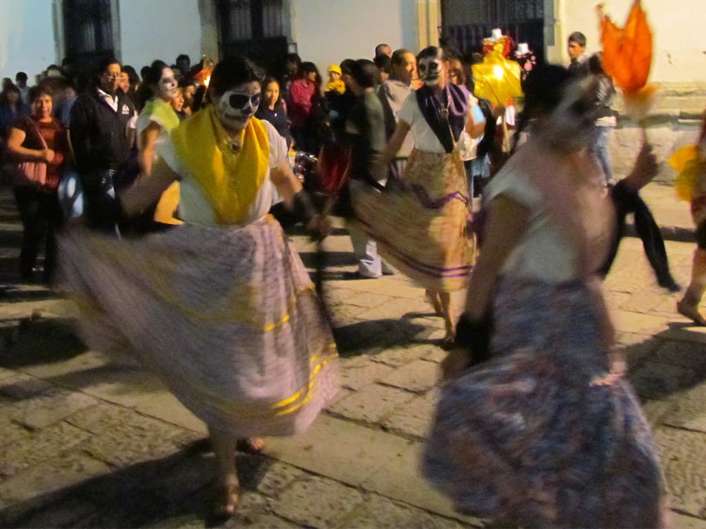 Dancers in skull face paint and full skirts swirl through a street comparsa in Oaxaca at night