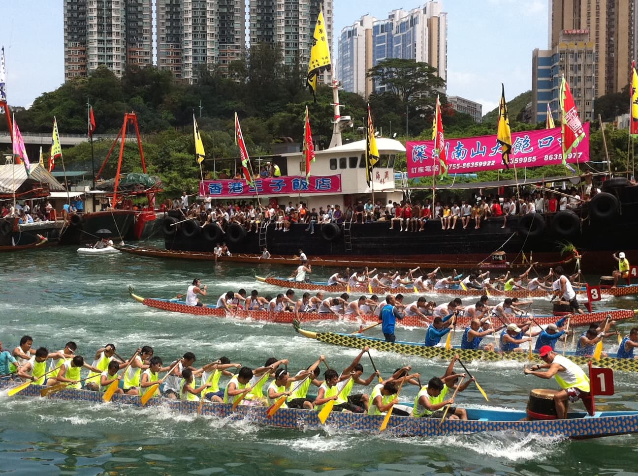 Crews in several dragon boats paddle hard in a race in a harbour lined with spectator boats and tower blocks