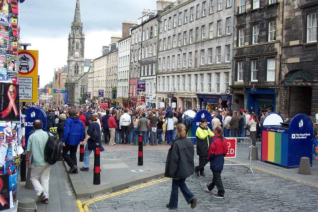 Crowds walk down Edinburgh's cobbled Royal Mile between stone tenements during the Fringe, with posters and street stalls along the sides