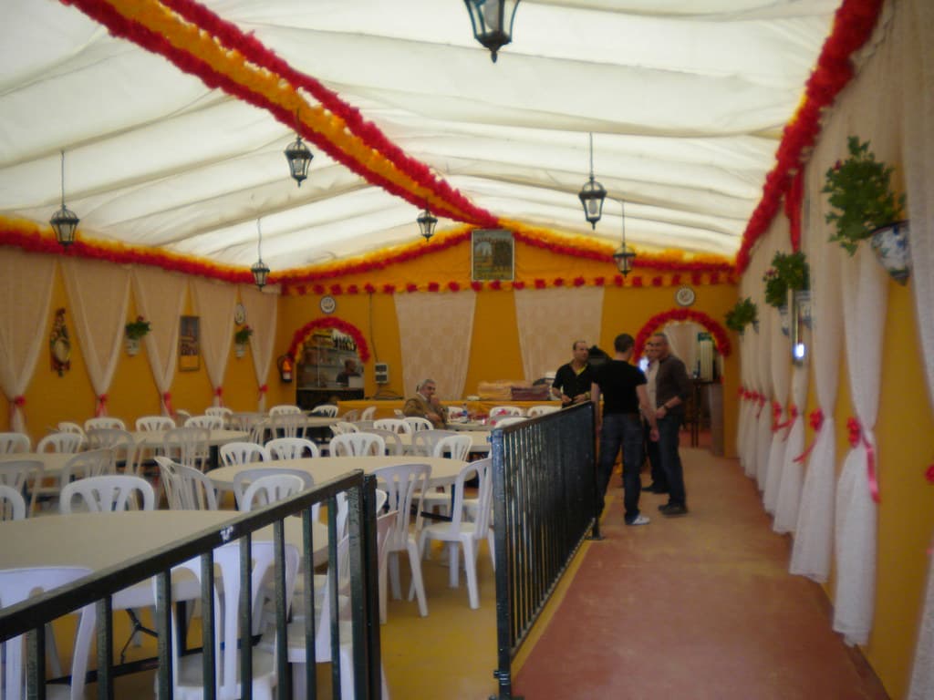 The inside of a caseta tent with yellow walls, white drapes and garlands of red and yellow paper flowers, with tables and chairs set out