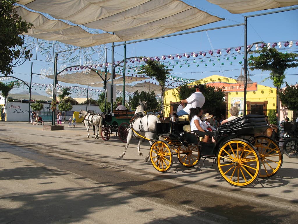 A horse-drawn carriage with yellow wheels and riders in traditional dress passes under decorated arches at the Málaga fair
