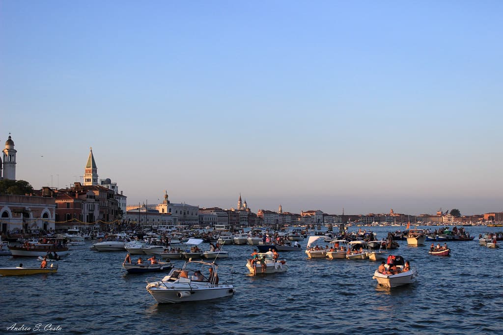 Dozens of small boats are moored across a calm lagoon at sunset, with the skyline and bell towers of Venice behind