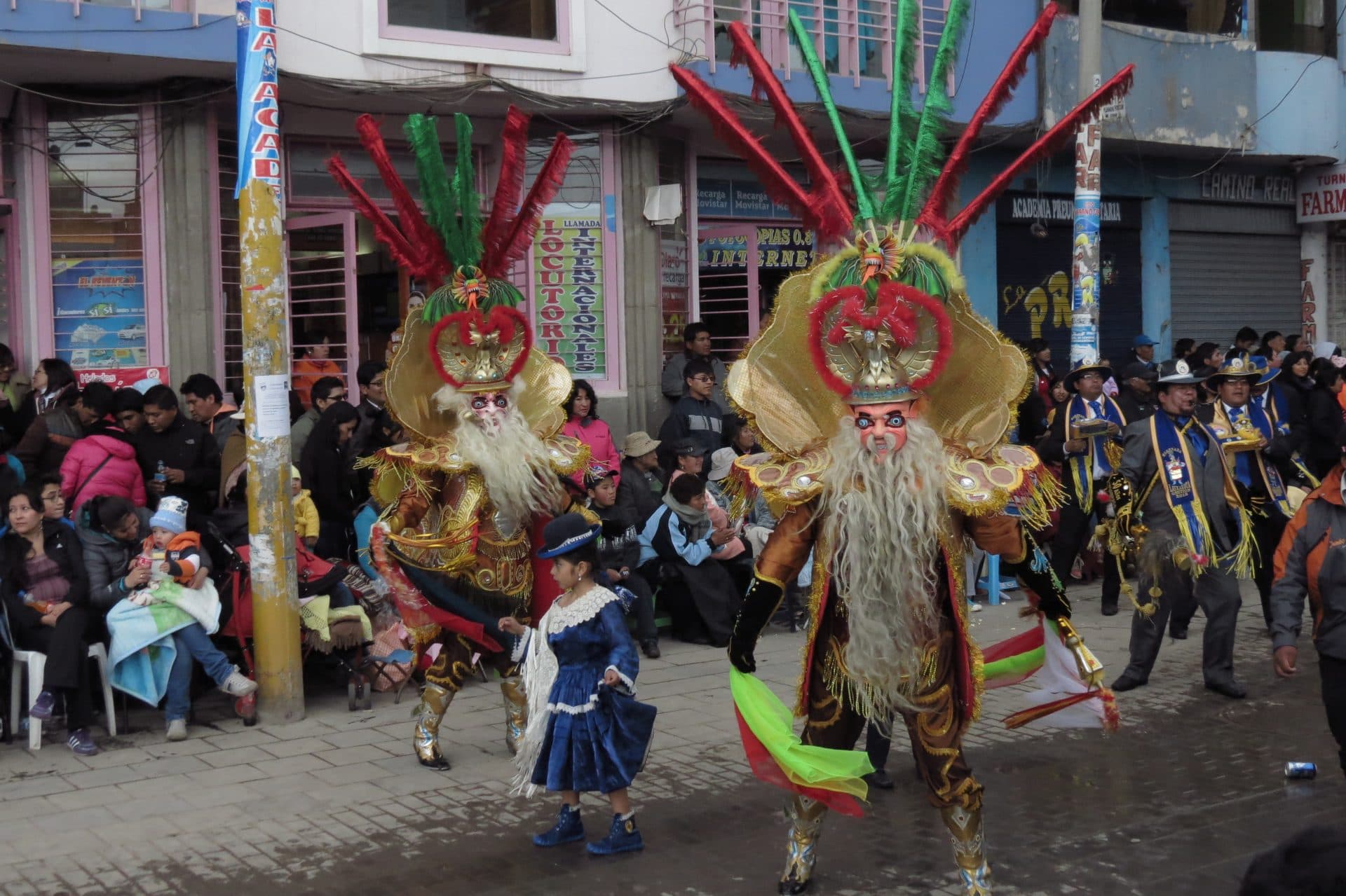 Two dancers in elaborate masks with long white beards, feathered headdresses and gold costumes move down a street while a small girl in a blue dress and a crowd look on