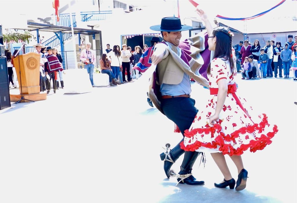 A couple dances the cueca, the man in a poncho and hat and the woman in a red and white dress, waving handkerchiefs