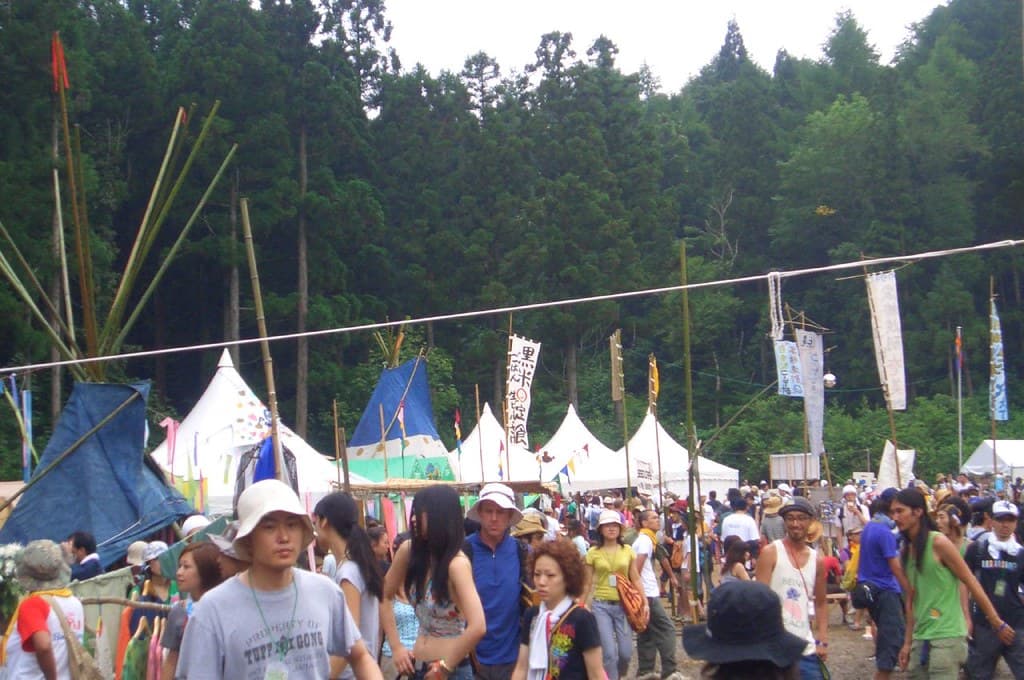 Festival-goers walk among white tents and flags under forested hills at Fuji Rock