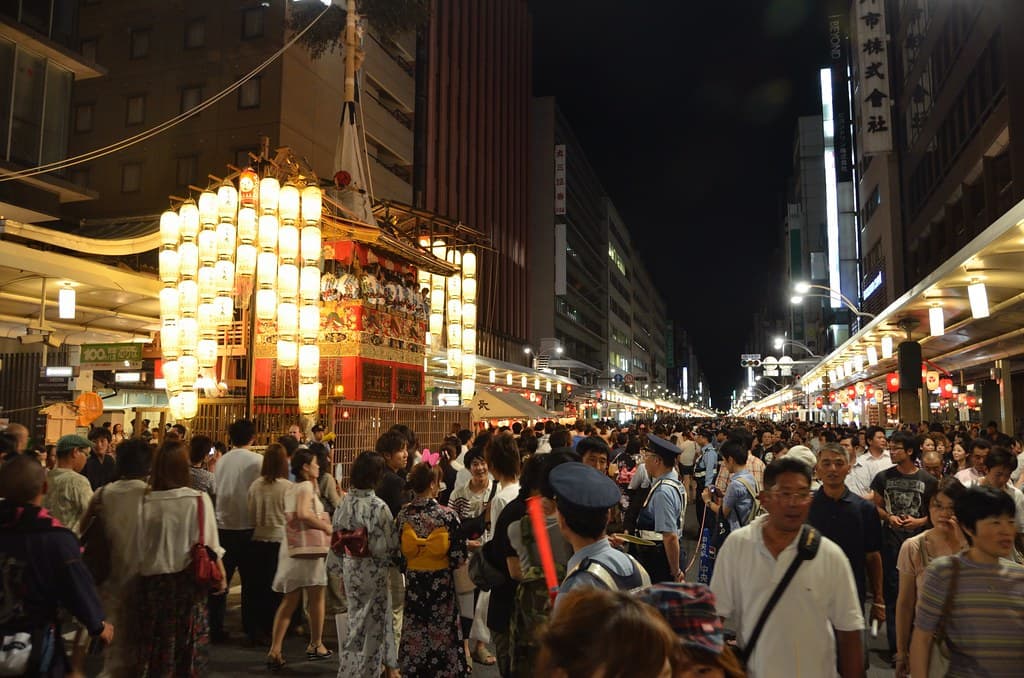 Crowds fill a Kyoto street at night, with a lantern-lit float glowing in the distance