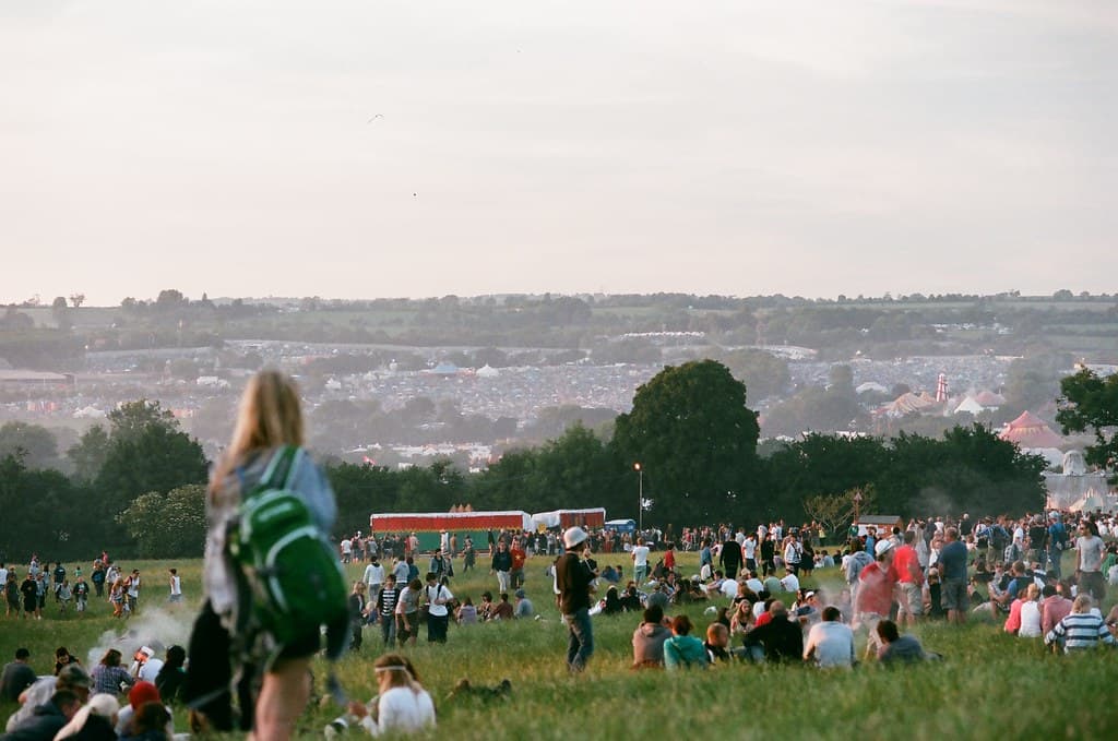 Festival-goers sit and walk on a grassy hillside at Glastonbury, with thousands of tents spread across the valley behind them