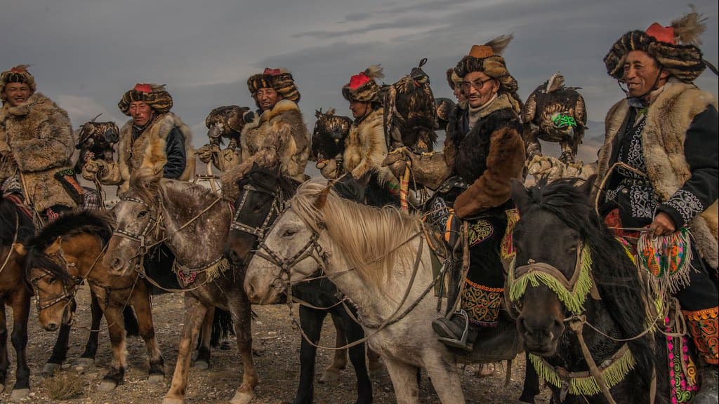 A line of Kazakh eagle hunters in fur hats and embroidered coats sit on their horses, golden eagles on their arms