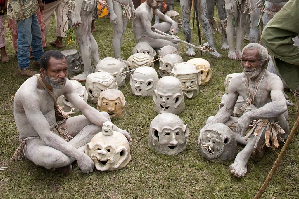 Asaro Mudmen covered in grey clay sit on the grass beside a row of large white clay masks at the Goroka Show