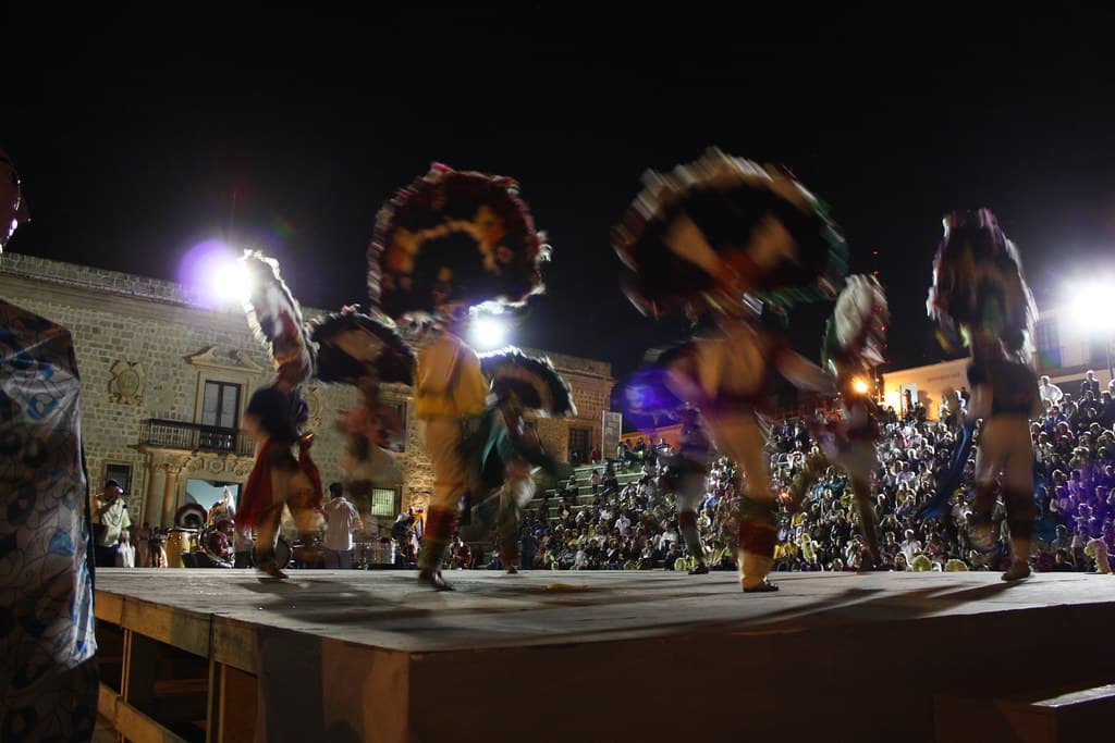 Danza de la Pluma dancers in huge round feathered headdresses whirl across an outdoor stage at night in front of a seated crowd