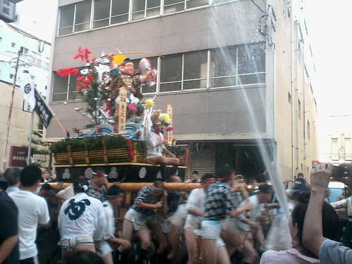 Men in happi coats and loincloths carry a decorated racing float through a Hakata street as spectators throw water over them