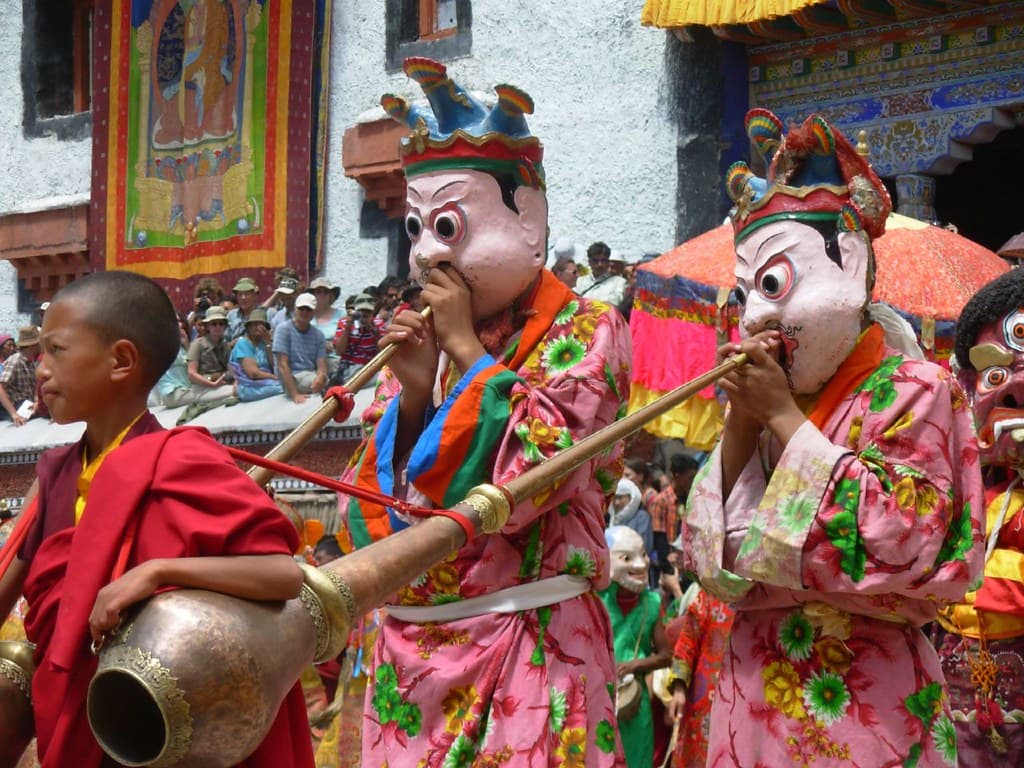 Masked dancers in pink and green brocade robes play long horns in the courtyard of Hemis Monastery, with a young monk beside them and crowds behind