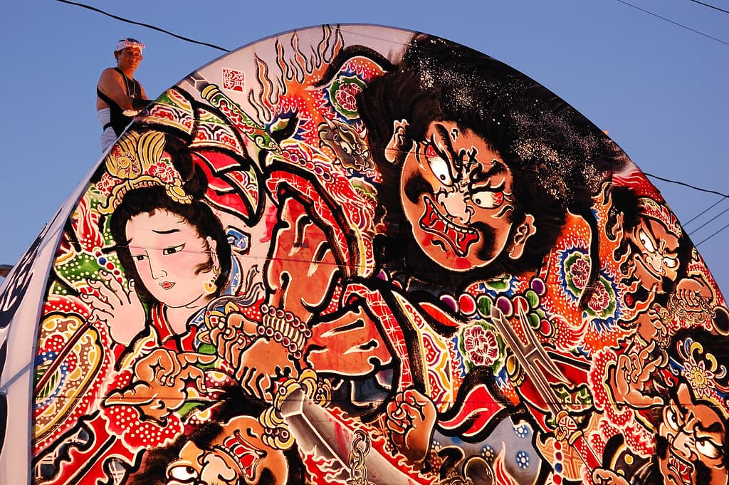 A painted fan-shaped neputa float showing fierce warriors in red and black, seen in daylight in Hirosaki