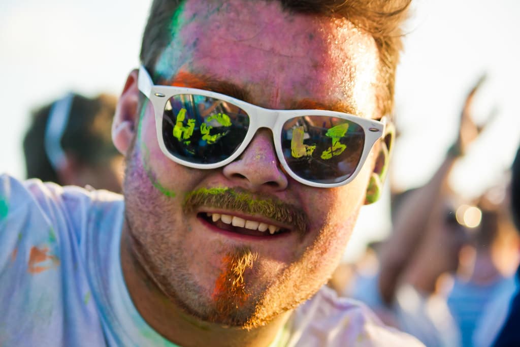 A smiling man in white sunglasses with colored powder on his face and hair
