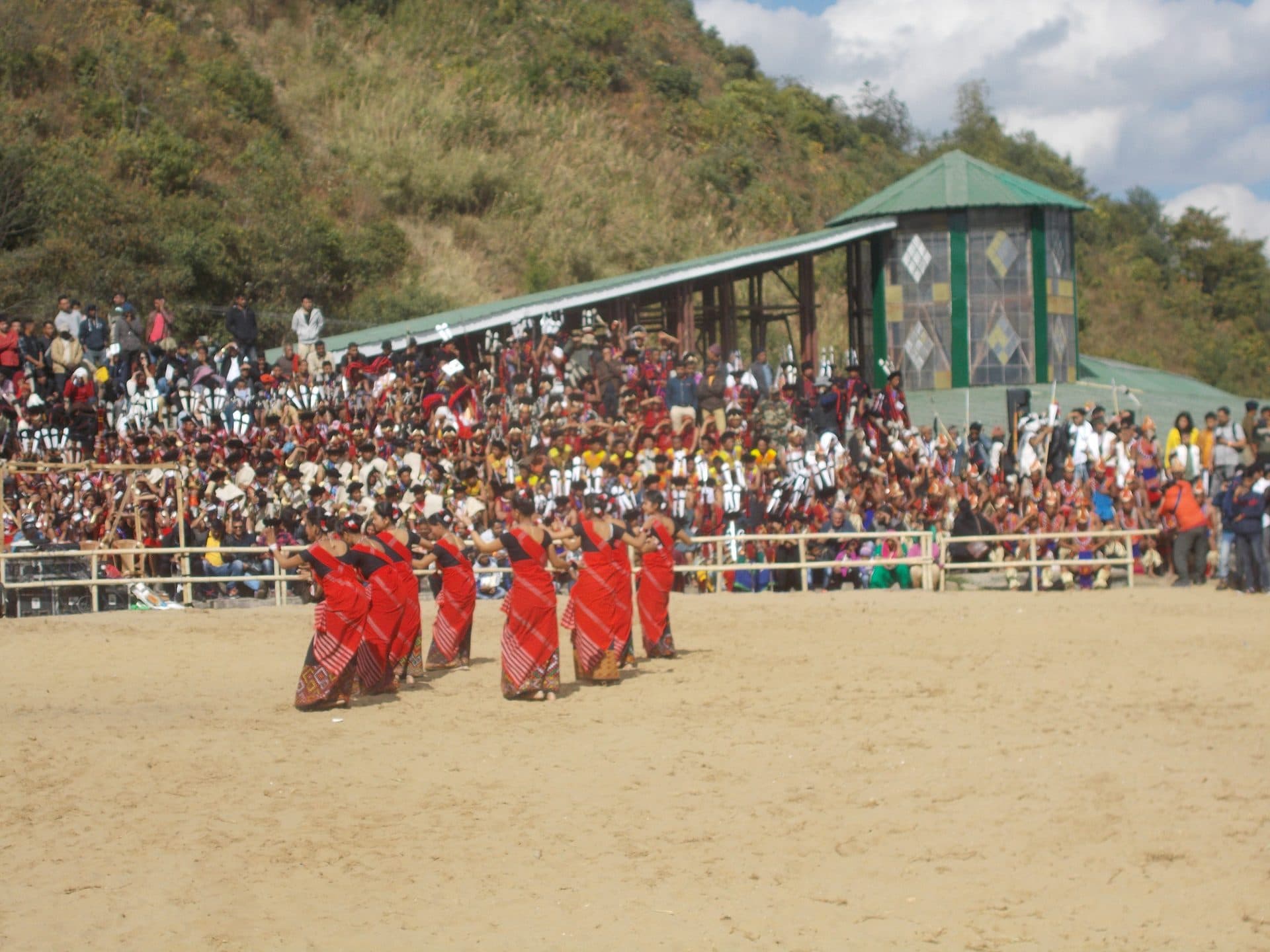 Women in red woven wraps dance in the arena before a crowd of participants in traditional Naga dress