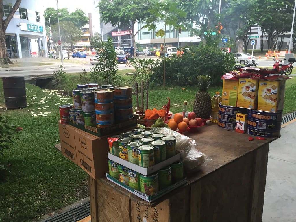 A table piled with canned food, biscuits, fruit and cooking oil set out as offerings on a patch of grass beside a street