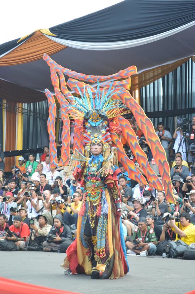 A performer in a huge feathered costume and headdress in blue, red and orange poses on the carpeted catwalk in front of a seated crowd at Jember Fashion Carnaval