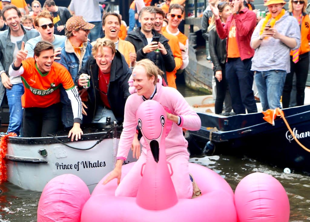 A man in a pink flamingo costume sits on an inflatable flamingo in a canal while cheerful people in orange watch from boats