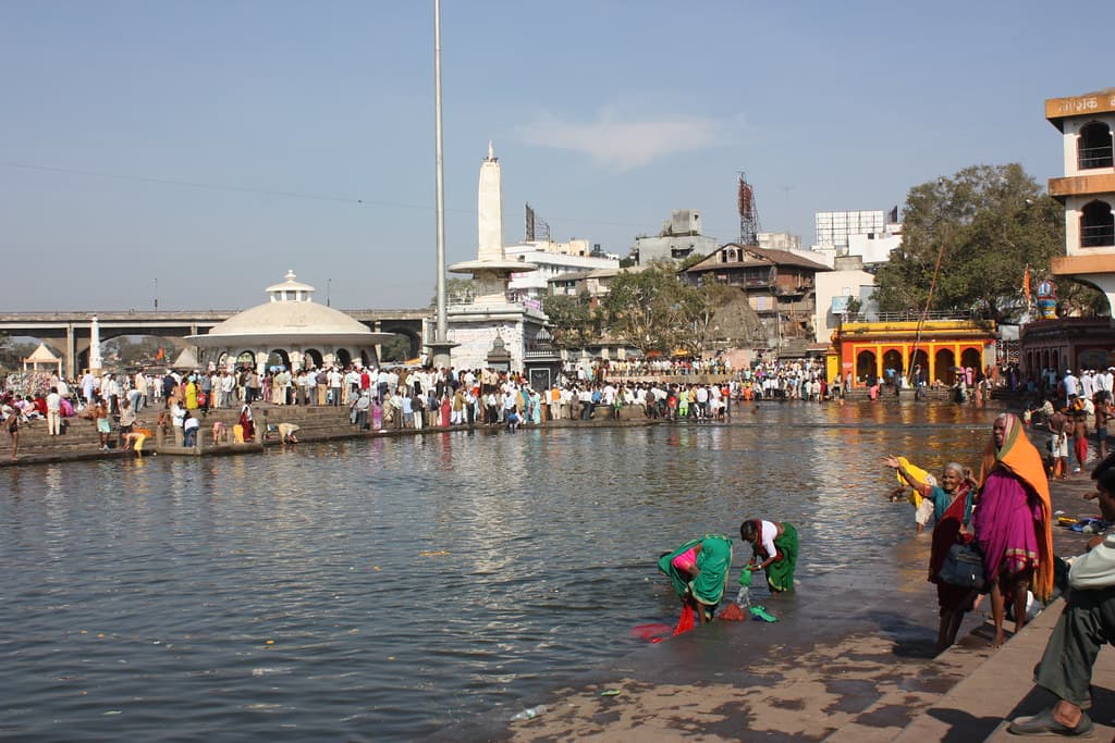 A pilgrim bathes at the edge of Ramkund on the Godavari in Nashik, with crowds along the far bank and temple domes behind