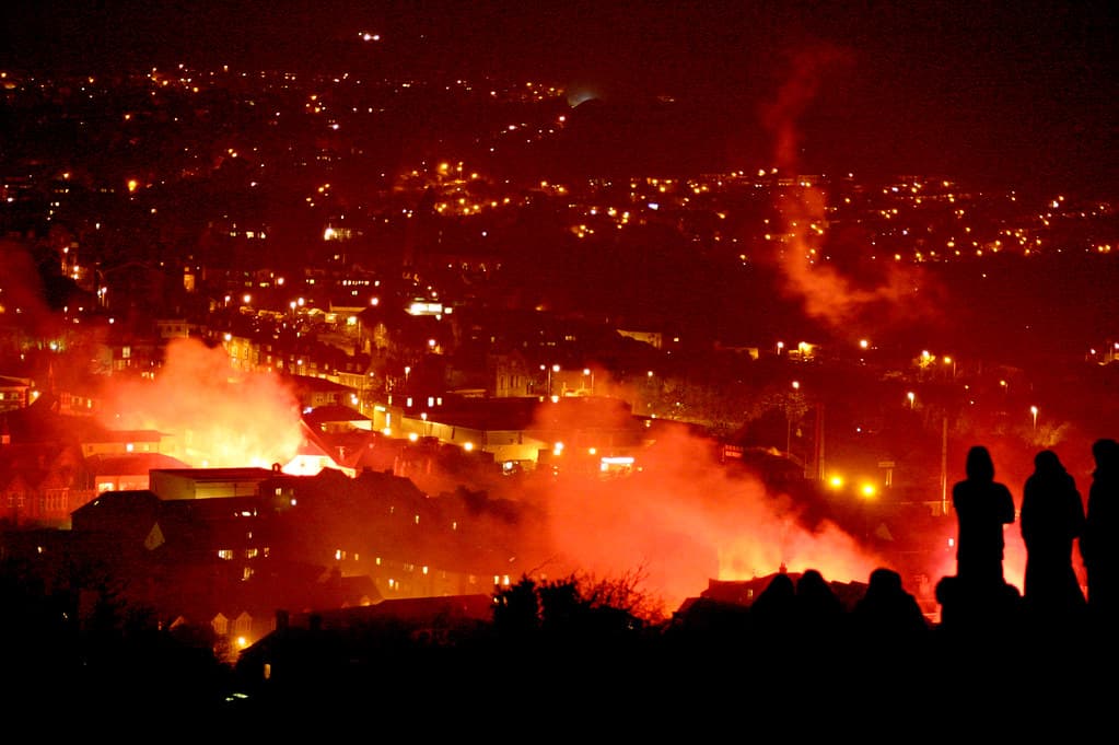 Silhouetted spectators on a hillside watch smoke and firelight rise over the town of Lewes