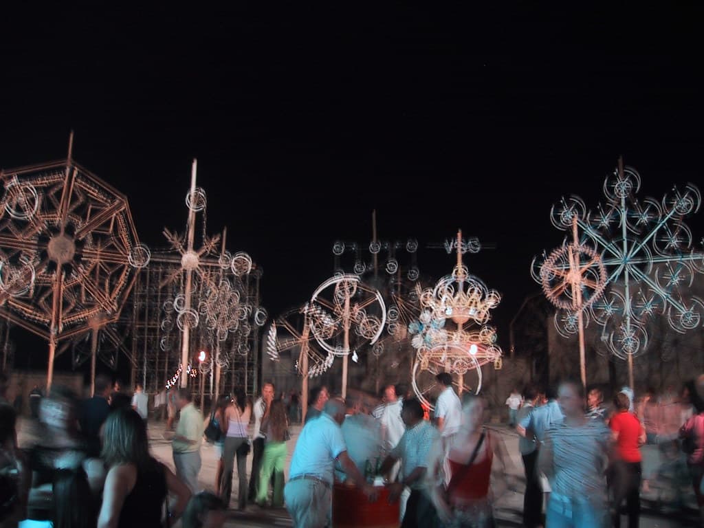 Crowds gather at night beneath tall ground fireworks set pieces in a Maltese village square during a festa