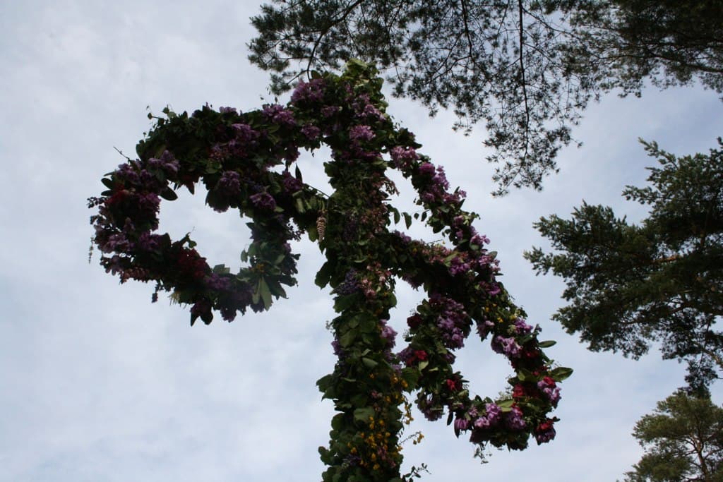 The cross-bar and hoops of a maypole wrapped thickly in leaves and purple flowers, seen from below against the sky and pine trees