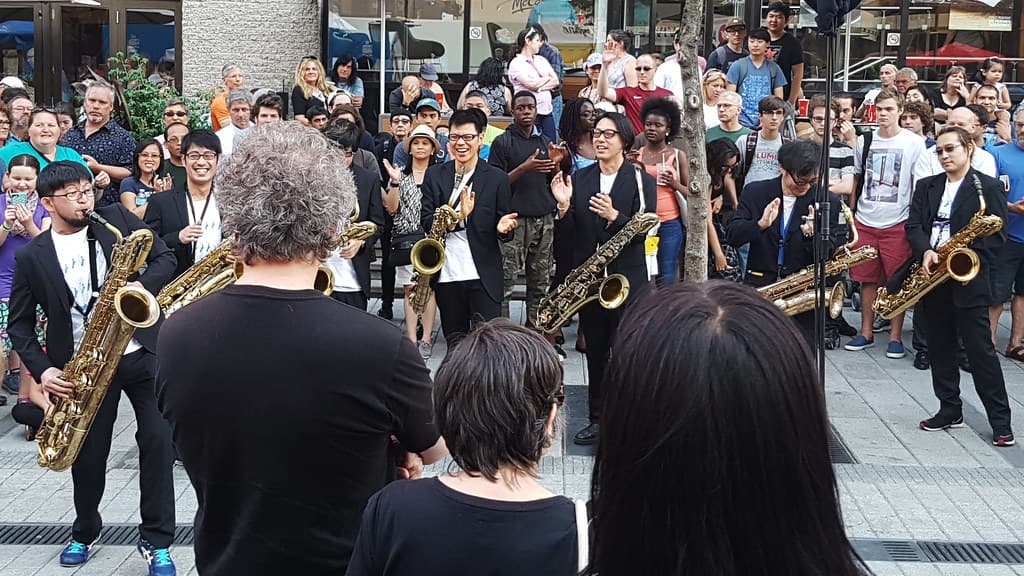 A brass band in dark suits plays saxophones and horns in the street surrounded by a crowd at the Montreal Jazz Festival