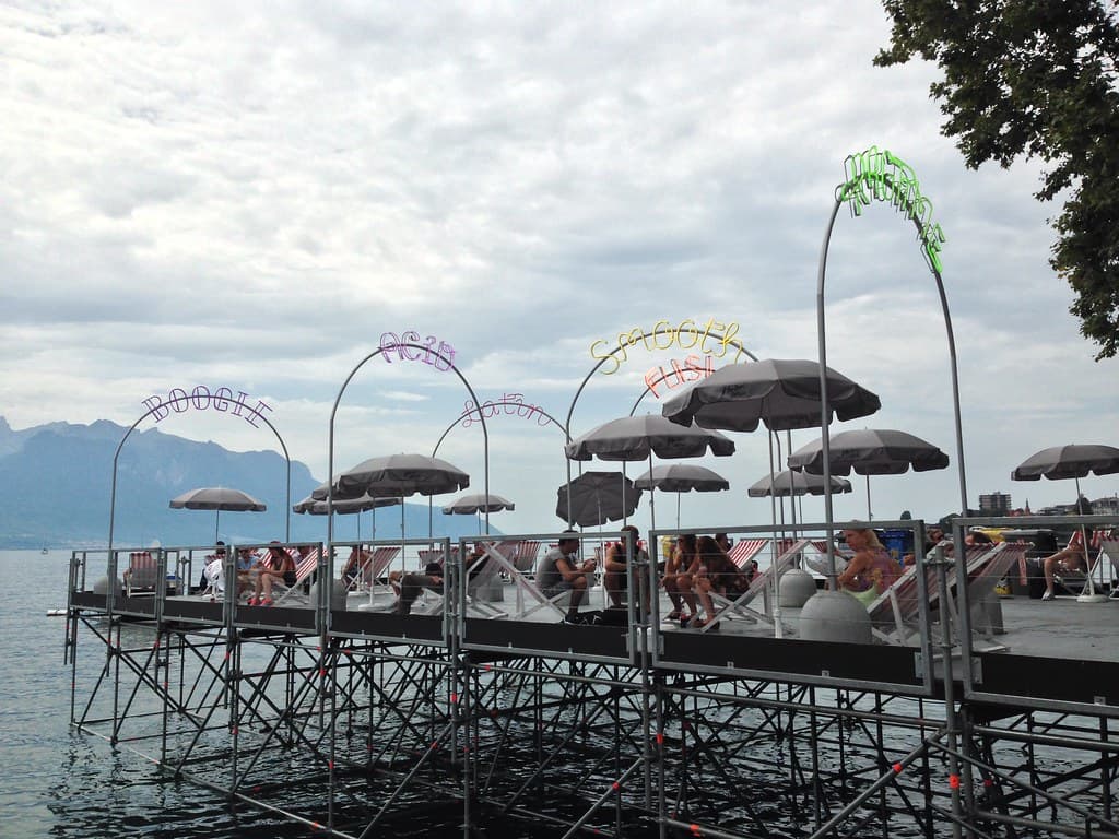 A wooden platform with umbrellas and illuminated arches stands over Lake Geneva at the Montreux Jazz Festival, with mountains across the water