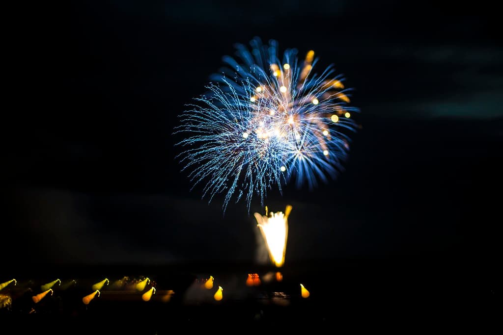 A blue and white firework bursts high over the dark Shinano River, with a line of lights along the far bank below