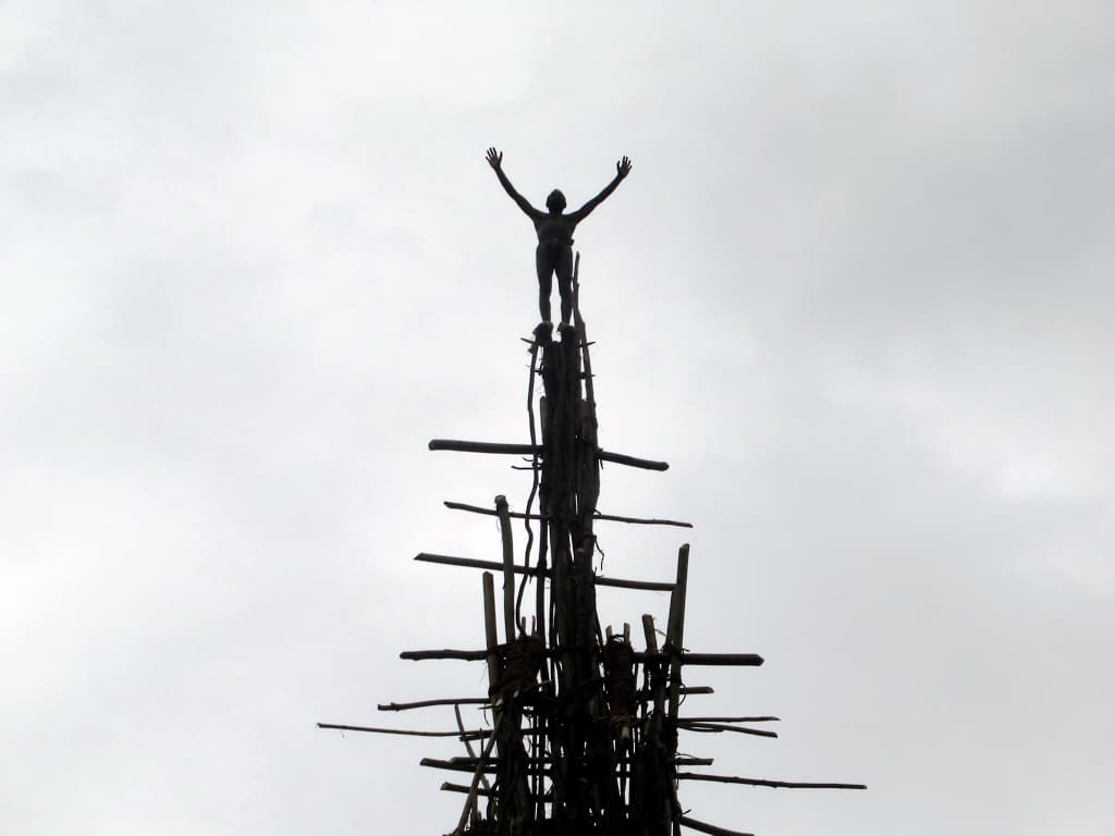 A land diver stands with arms outstretched on top of a tall wooden tower against a grey sky