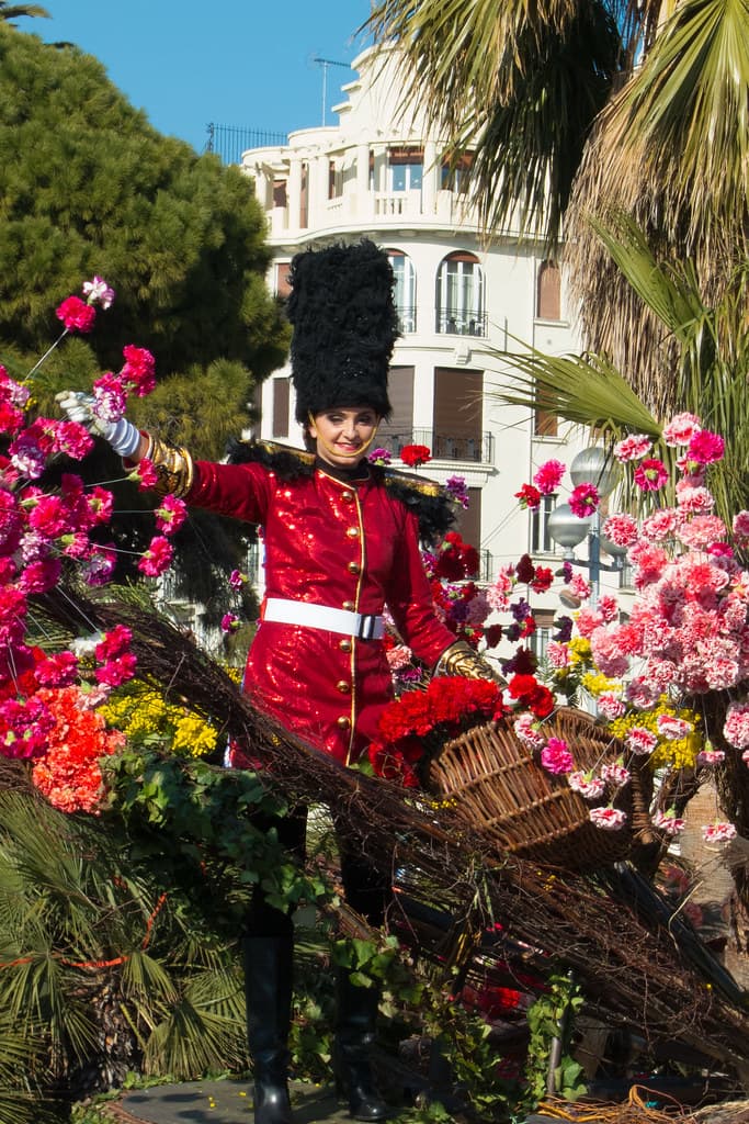 A performer in a red guardsman's costume stands on a float covered in pink and red flowers at a Nice flower battle