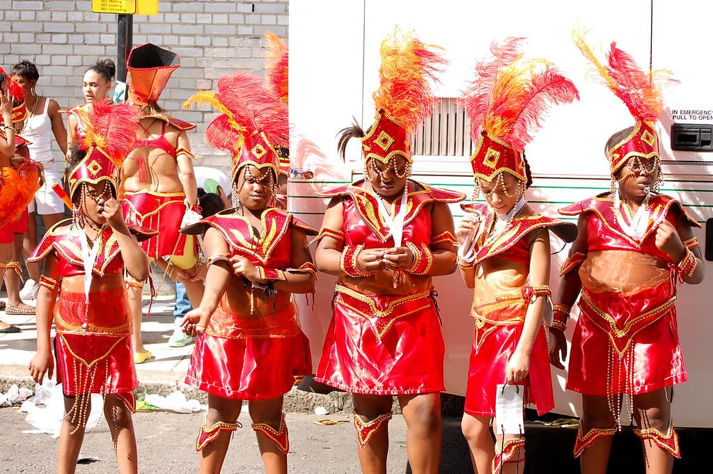 Young masqueraders in matching red sequinned costumes and feathered headdresses stand together on a street during Notting Hill Carnival