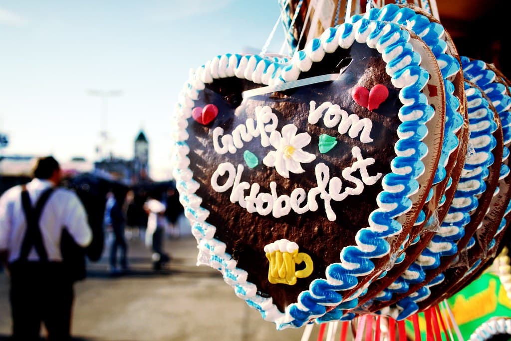 Gingerbread hearts iced with Gruss vom Oktoberfest hang at a stall, with fairgoers in traditional dress walking past