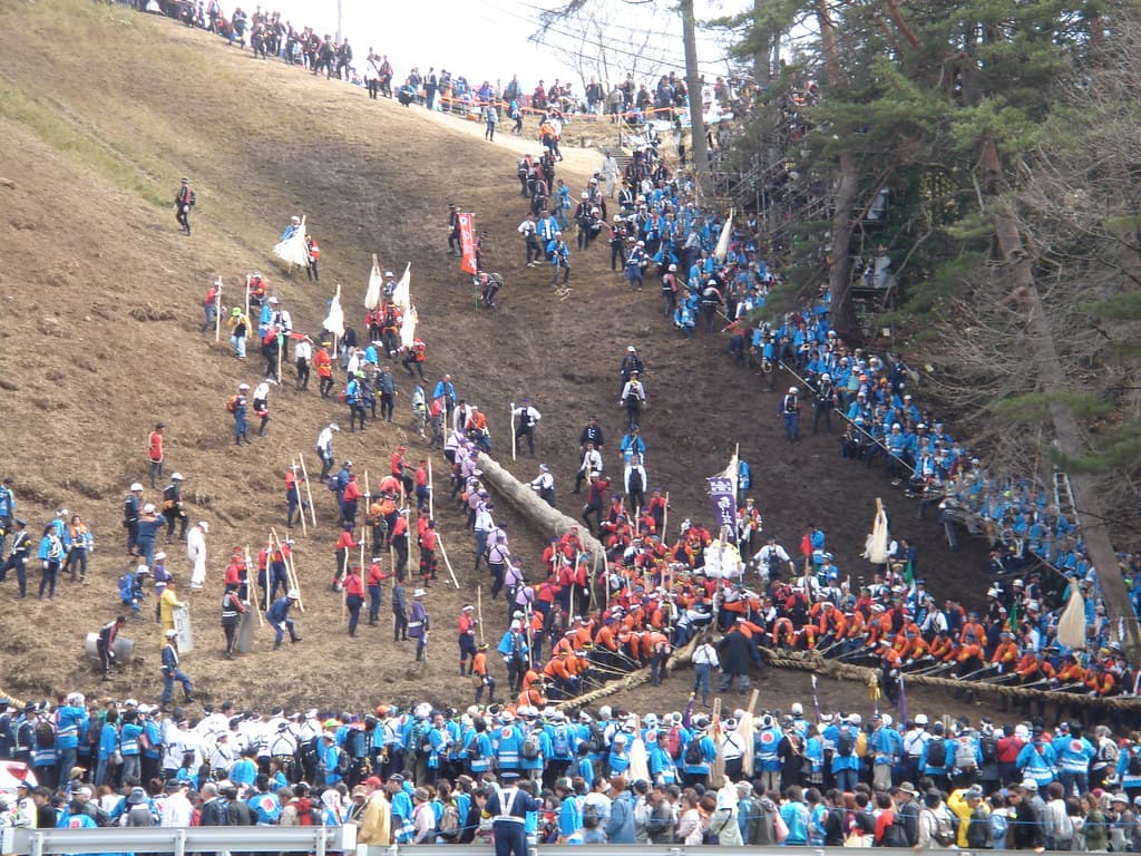 Crowds in blue and red happi coats haul a huge log down a steep bare hillside