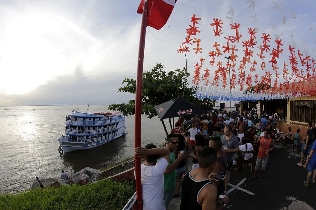 Crowds gather on the Parintins waterfront under strings of red Boi Garantido decorations, with a riverboat moored on the Amazon