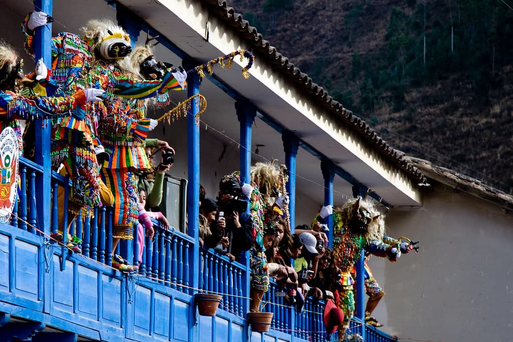 Masked dancers in colourful costumes crowd a blue wooden balcony above a street in Paucartambo during the fiesta