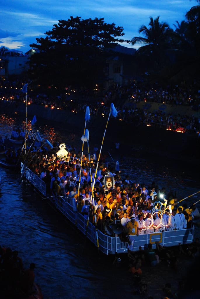 A decorated barge crowded with devotees and candles moves along the Naga River at dusk during the Peñafrancia fluvial procession
