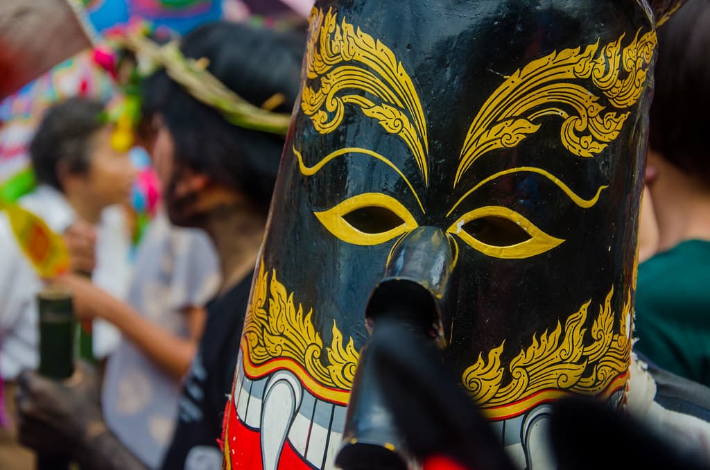 A black, gold and red Phi Ta Khon mask with a long nose seen up close in a festival crowd, with children behind