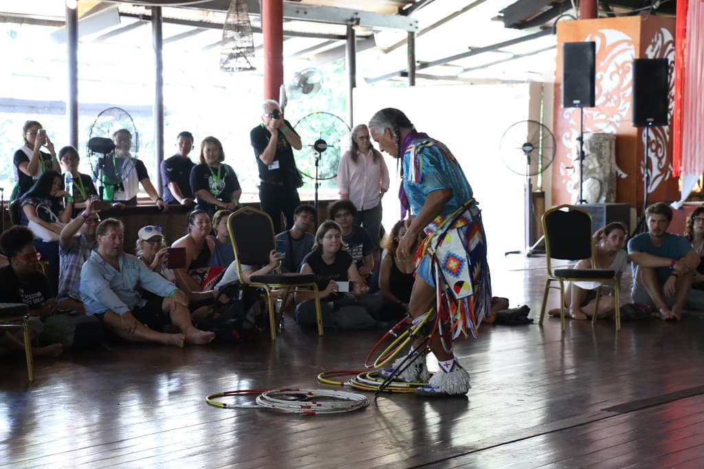 An audience sits on the wooden floor of an open-sided hall watching a performer in Native American regalia lead a hoop dance workshop