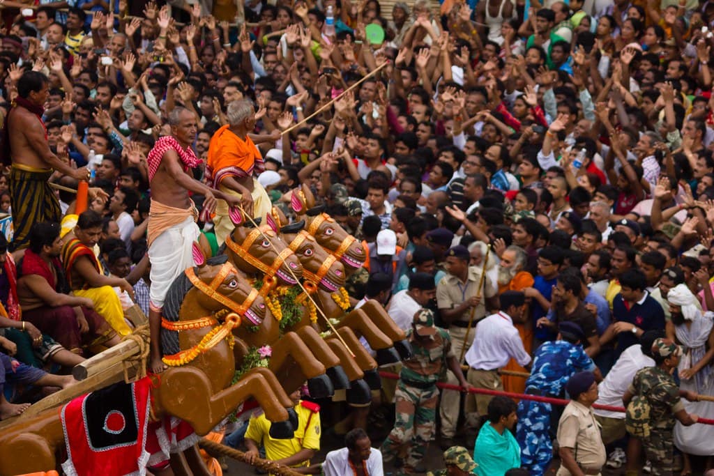 A dense crowd of devotees surrounds the base of a decorated wooden temple chariot during Rath Yatra