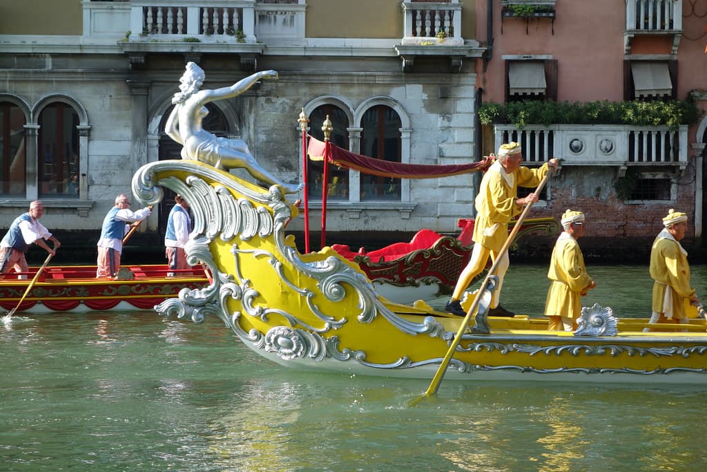 Rowers in yellow costumes row an ornate yellow boat with a carved silver prow along the Grand Canal in Venice