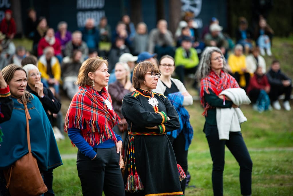 Festival-goers in Sámi dress and red shawls stand in the audience at an outdoor event at Riddu Riđđu in Manndalen