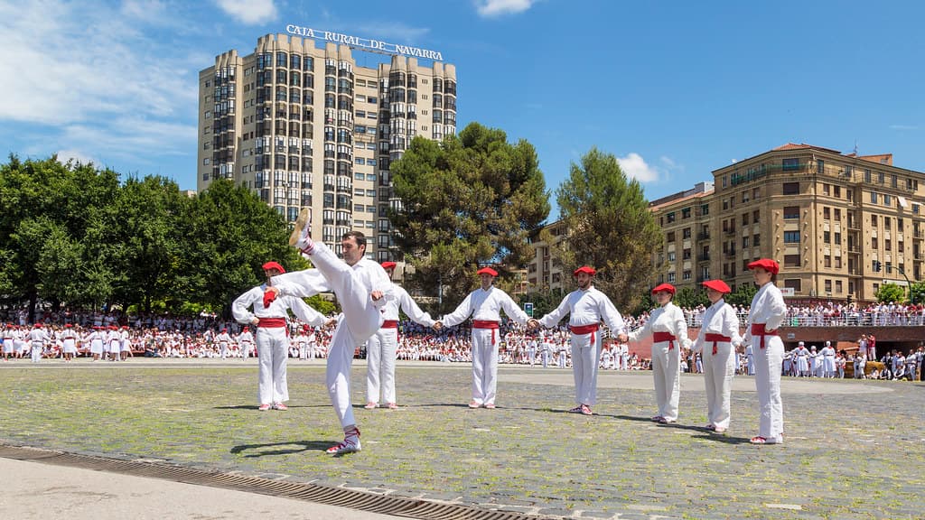 Dancers in white with red sashes and neckerchiefs perform in a line on a sunny square in Pamplona