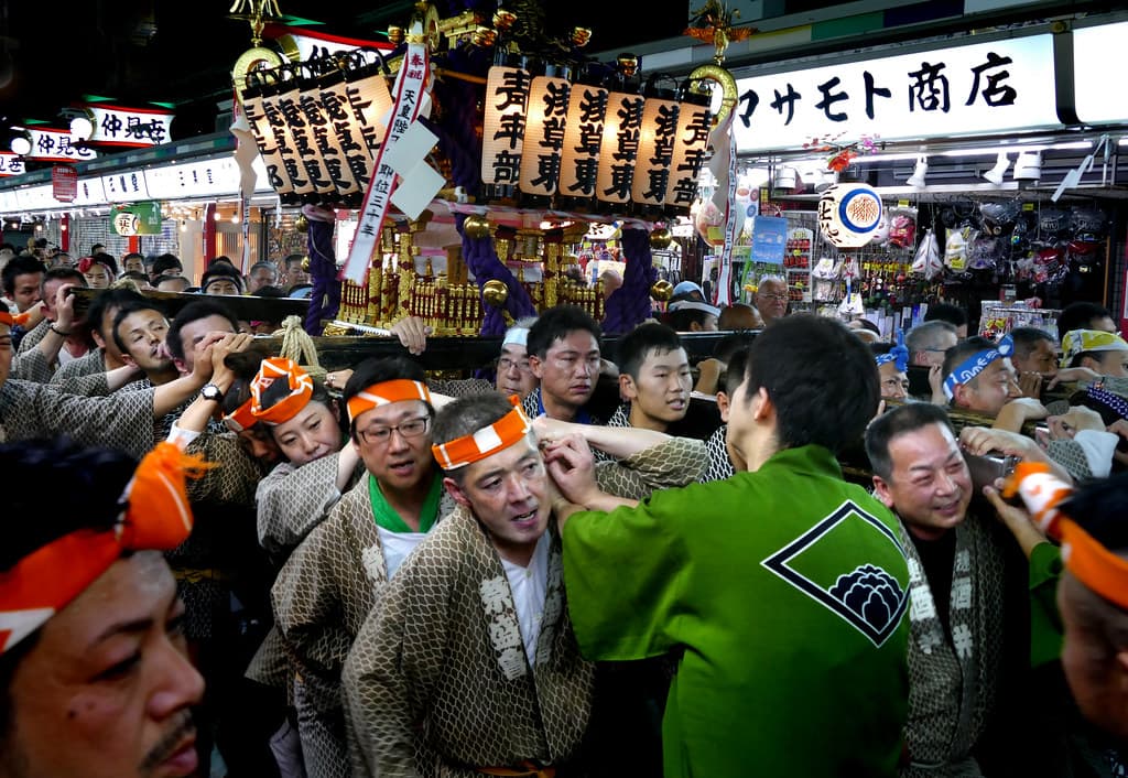 A crowd in festival headbands and coats pushes a gold and black portable shrine past shops in Asakusa