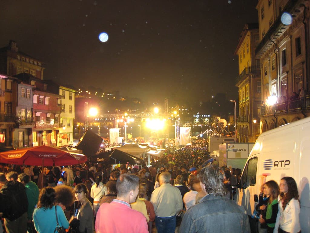 A dense crowd fills Porto's riverside Ribeira square at night during São João, with stalls, umbrellas and a lit stage in the distance