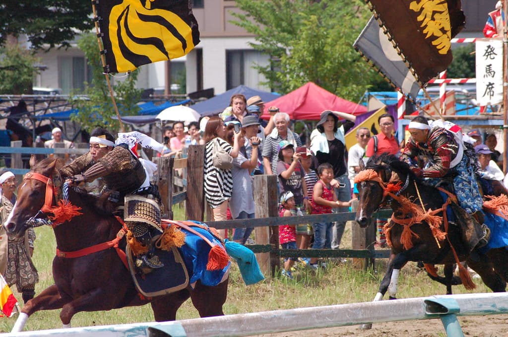 Two armoured riders with banners on their backs gallop side by side past a crowd of spectators along a fenced dirt track